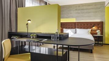 Habitación de hotel con cama, escritorio y silla, lámpara, mesita con un florero y mesita de noche con una flor.