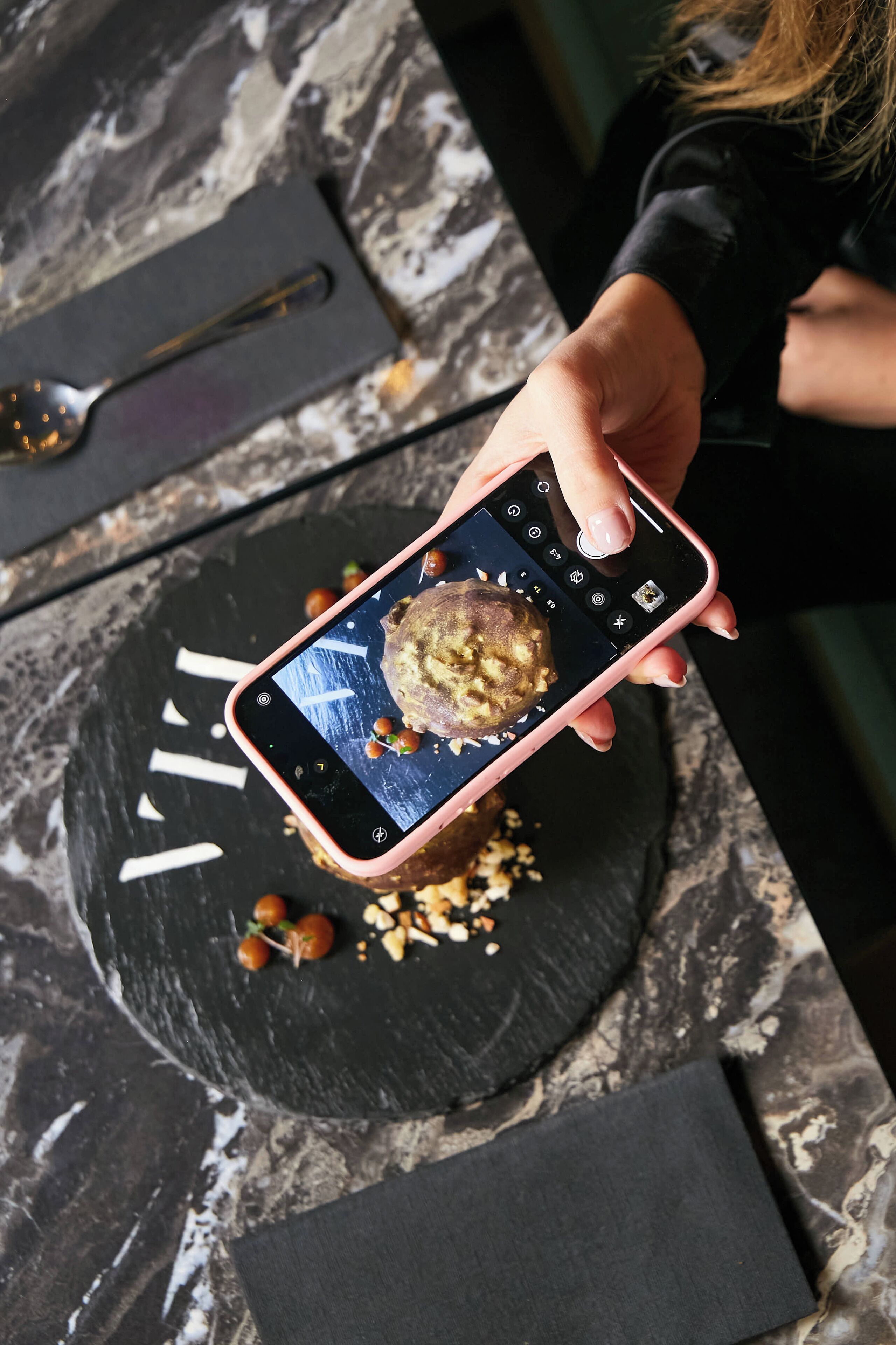 Eine Person hält ein Smartphone, auf einem schwarzen Schieferteller liegt ein Dessert.