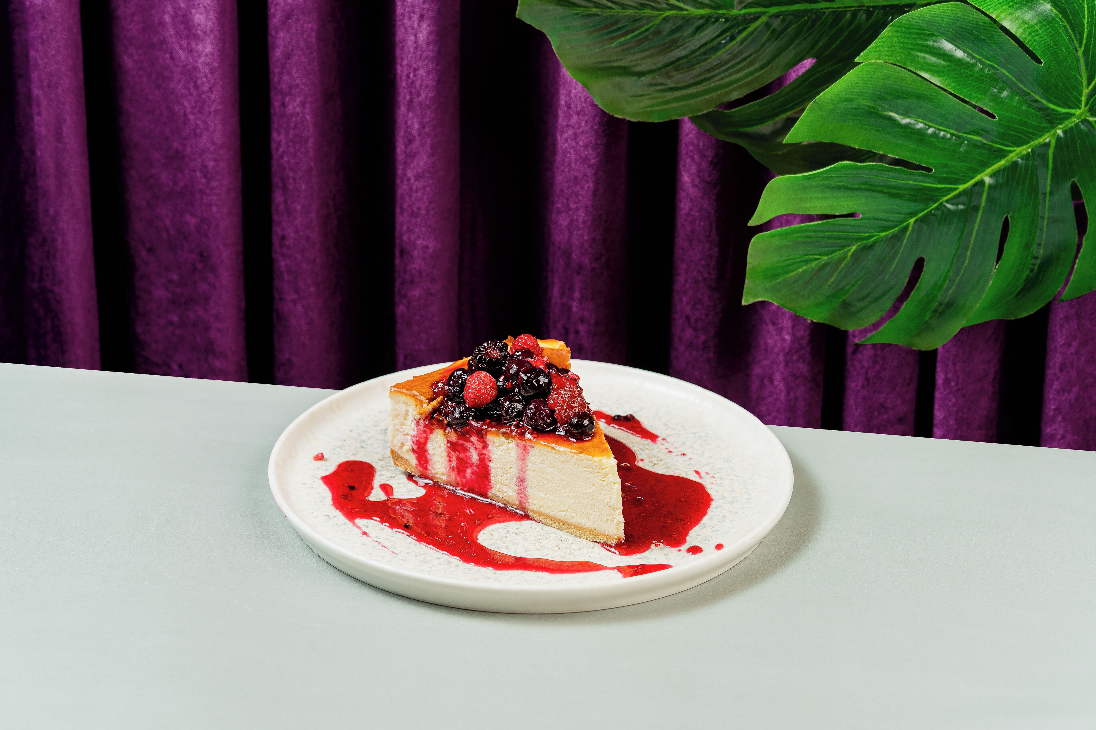 Una porción de pastel de queso en un plato blanco con salsa de frutos rojos, sobre un fondo morado.
