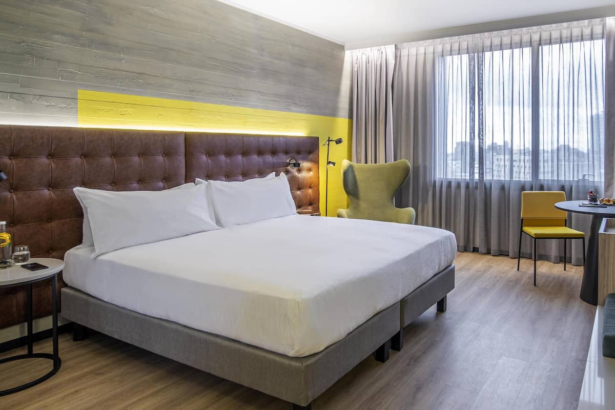 Ein Hotelzimmer mit einem großen Bett, einem Schreibtisch mit Stuhl, einer Lampe und Blick auf die Stadt durch das Fenster.