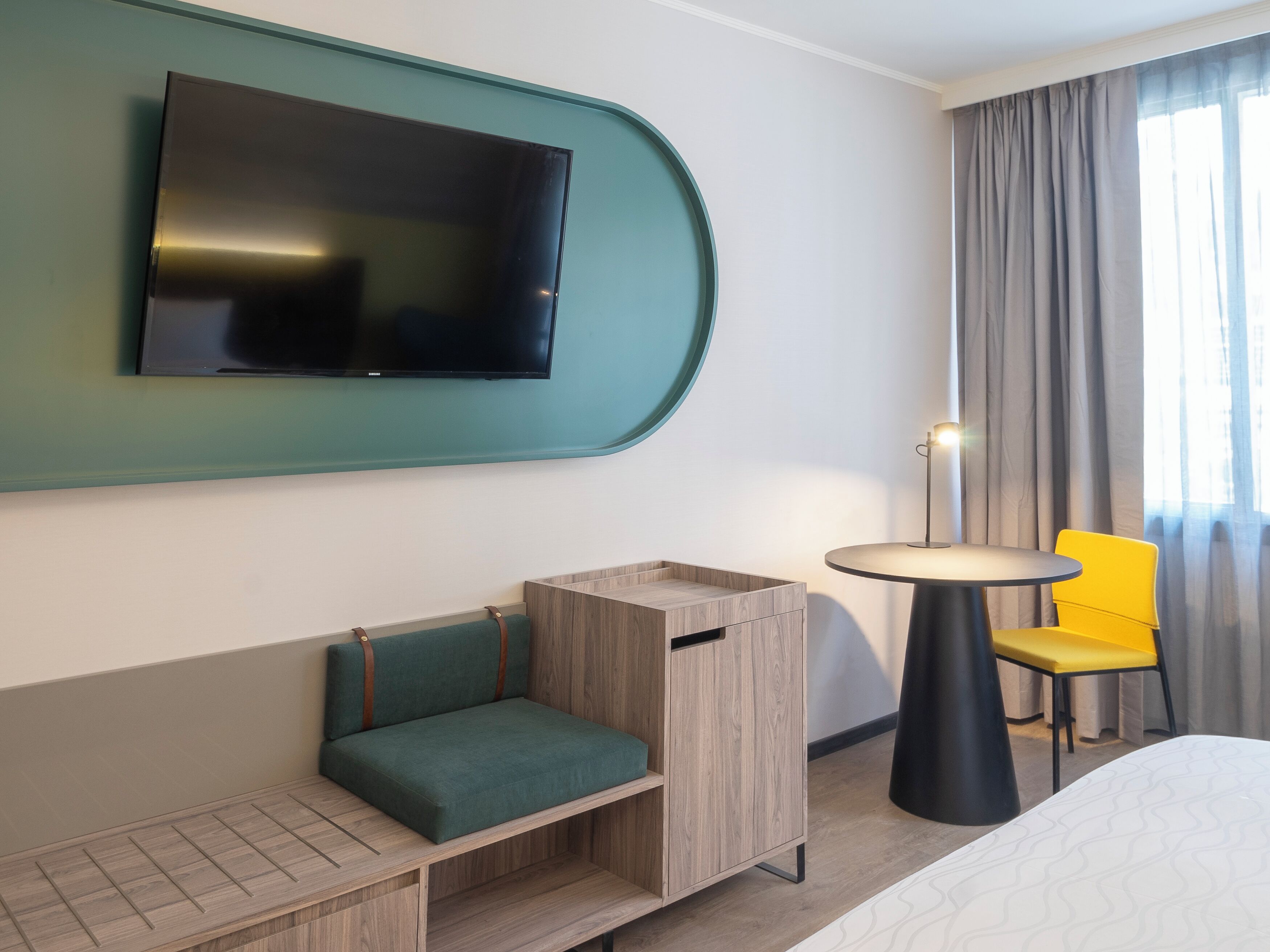 Hotelkamer met een flatscreen televisie bevestigd aan een blauwgroene muur, een klein houten kastje, een groene met kussens beklede bank, een ronde tafel met een gele stoel en een bed met een beddegoed met een patroon.