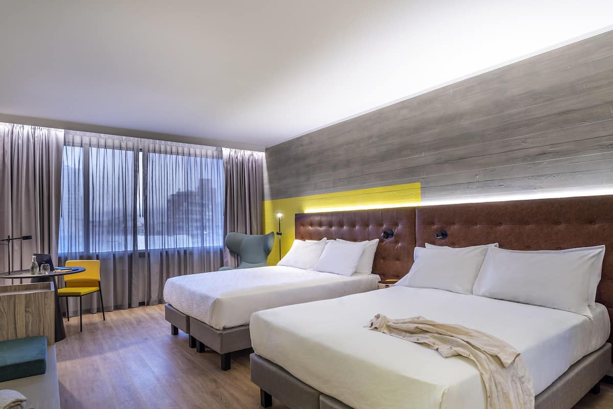 A hotel room with two beds, a desk with a chair, a window with curtains, and a modern headboard.