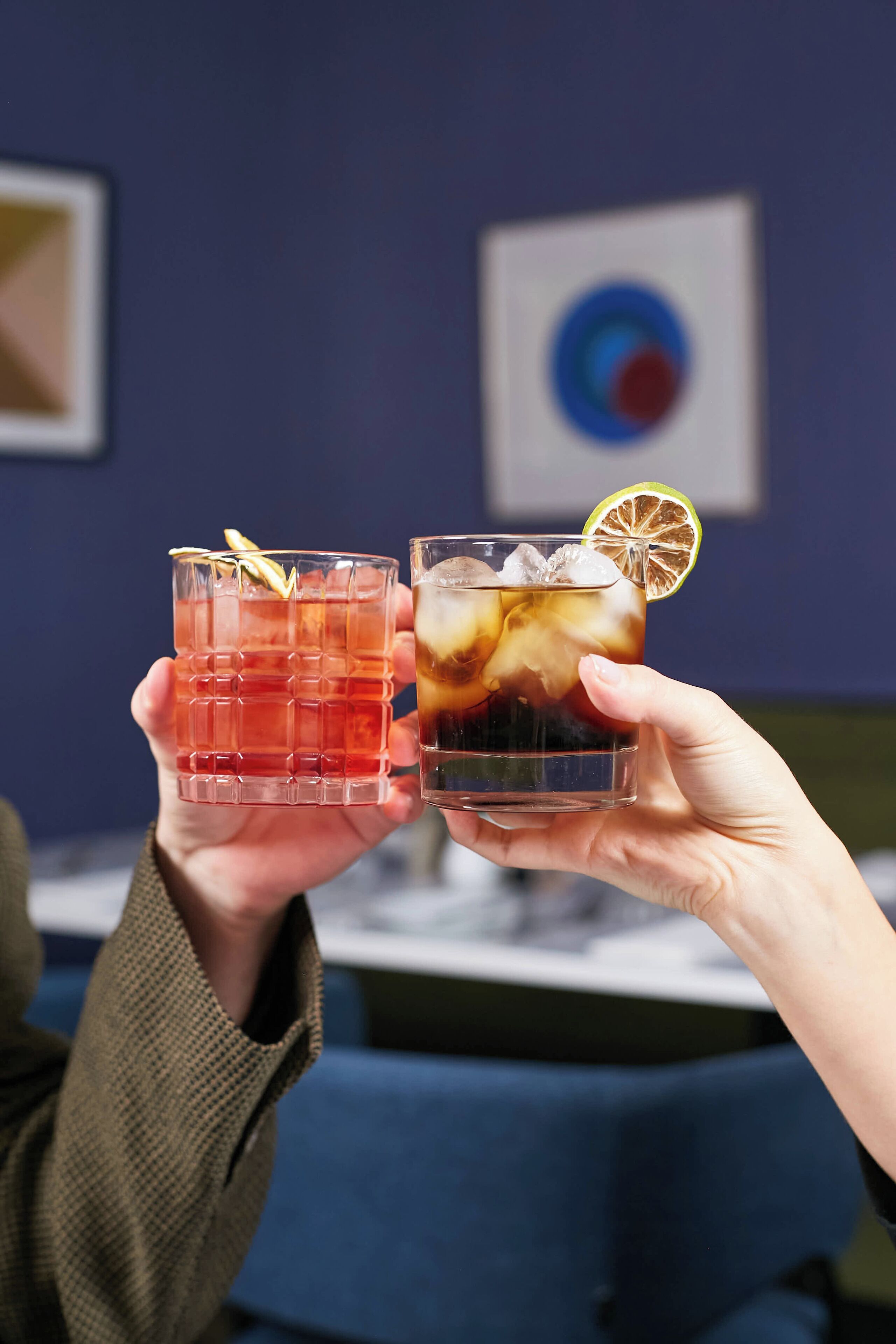 Two hands holding two glasses of iced drinks, one with a lime slice garnish.