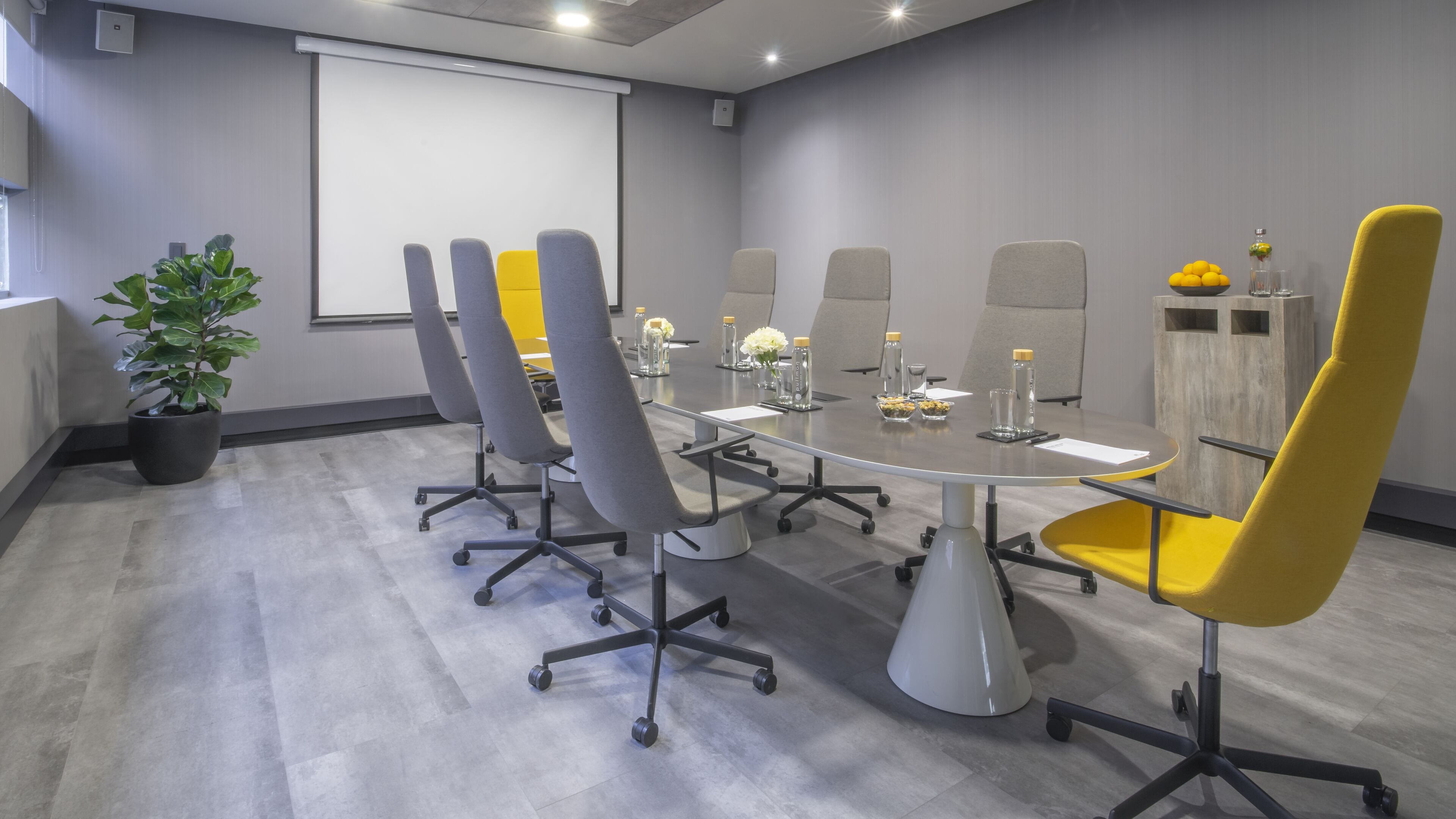 A conference room with a large table, six chairs, a projector screen, and a potted plant.