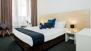 A hotel room with a large bed, a bedside table with a lamp, a chair, and a window with curtains. - Nomads Sydney - Hostel (Sydney)