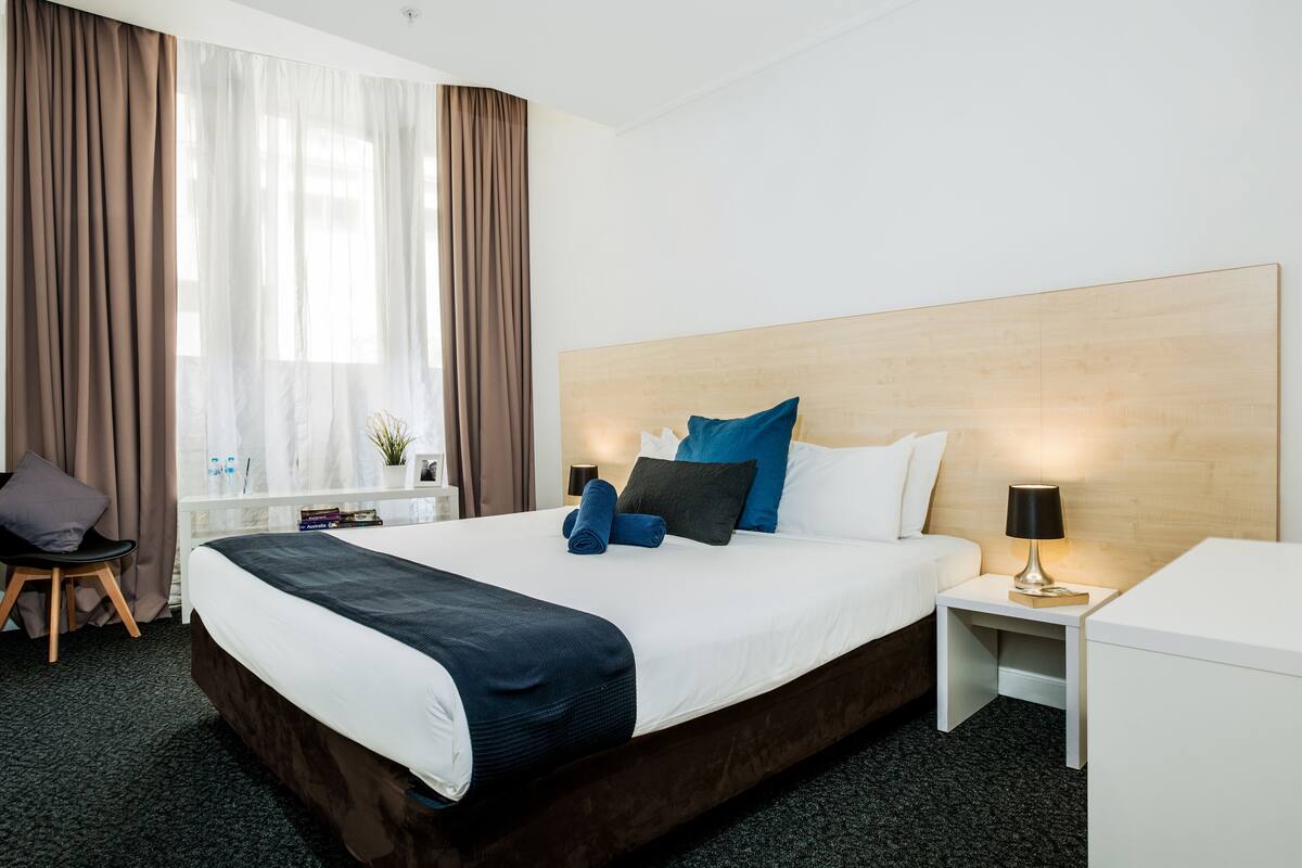 A hotel room with a large bed, a bedside table with a lamp, a chair, and a window with curtains.