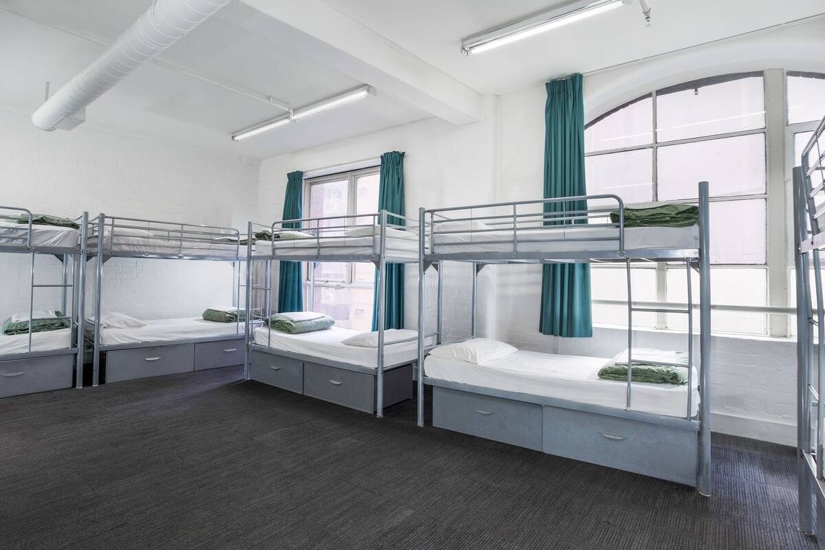A dormitory room with bunk beds, a window with curtains, and a staircase.