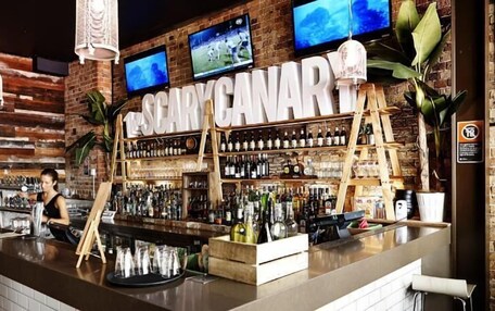 A bar with a rustic brick wall, shelves of liquor bottles, and two TVs mounted above the bar area.