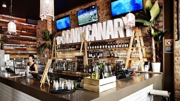 A bar with a rustic brick wall, shelves of liquor bottles, and two TVs mounted above the bar area. - Nomads Sydney - Hostel (Sydney)