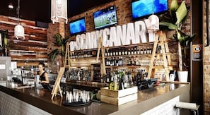 A bar with a rustic brick wall, shelves of liquor bottles, and two TVs mounted above the bar area. - Nomads Sydney - Hostel (Sydney)