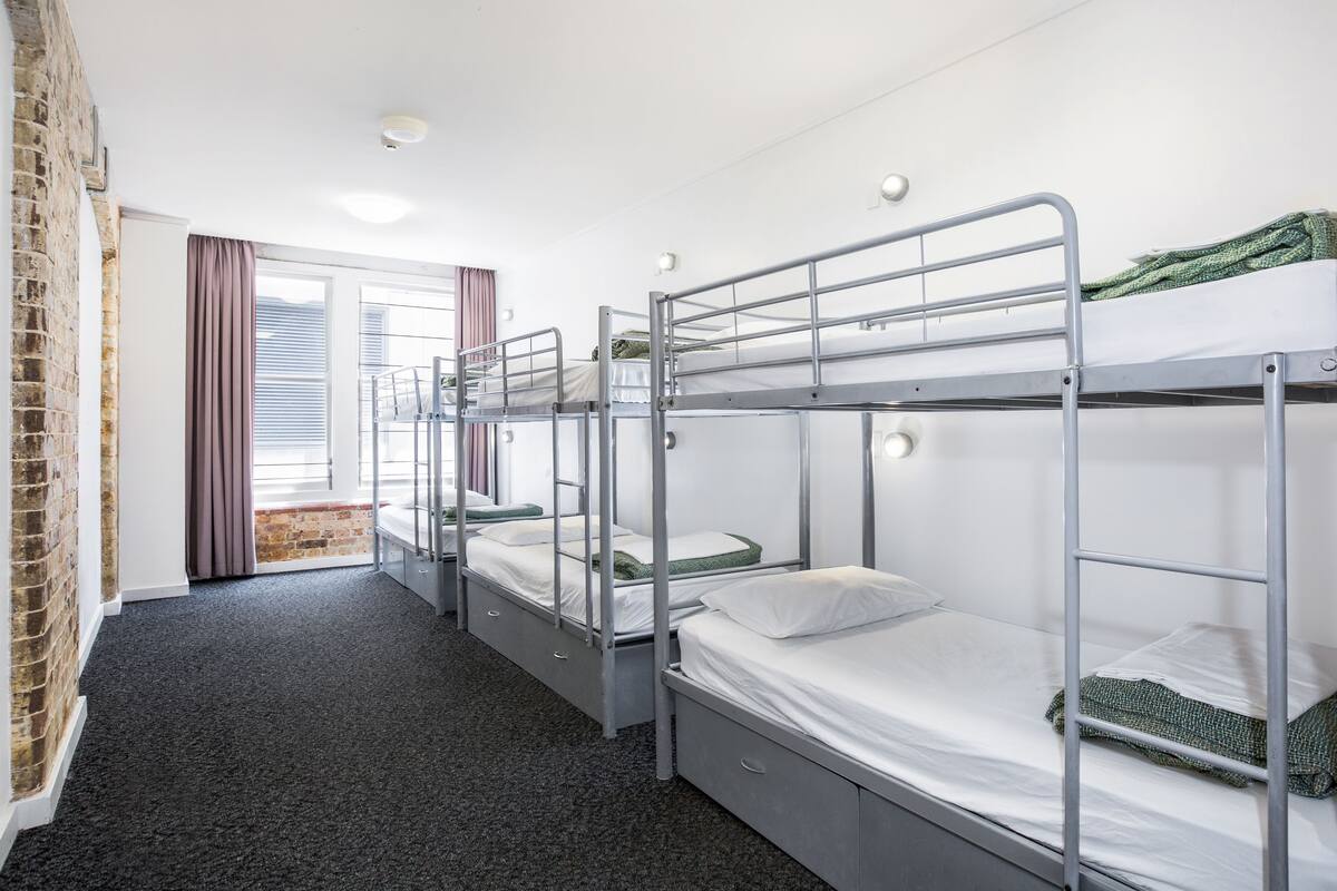 A dormitory room with bunk beds, a brick wall, and a window with curtains.