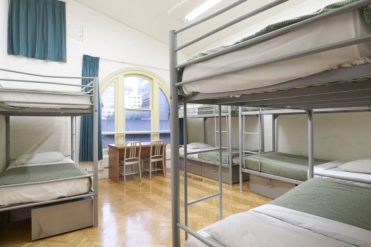 A dormitory room with bunk beds, a wooden floor, a desk, and a window with curtains.