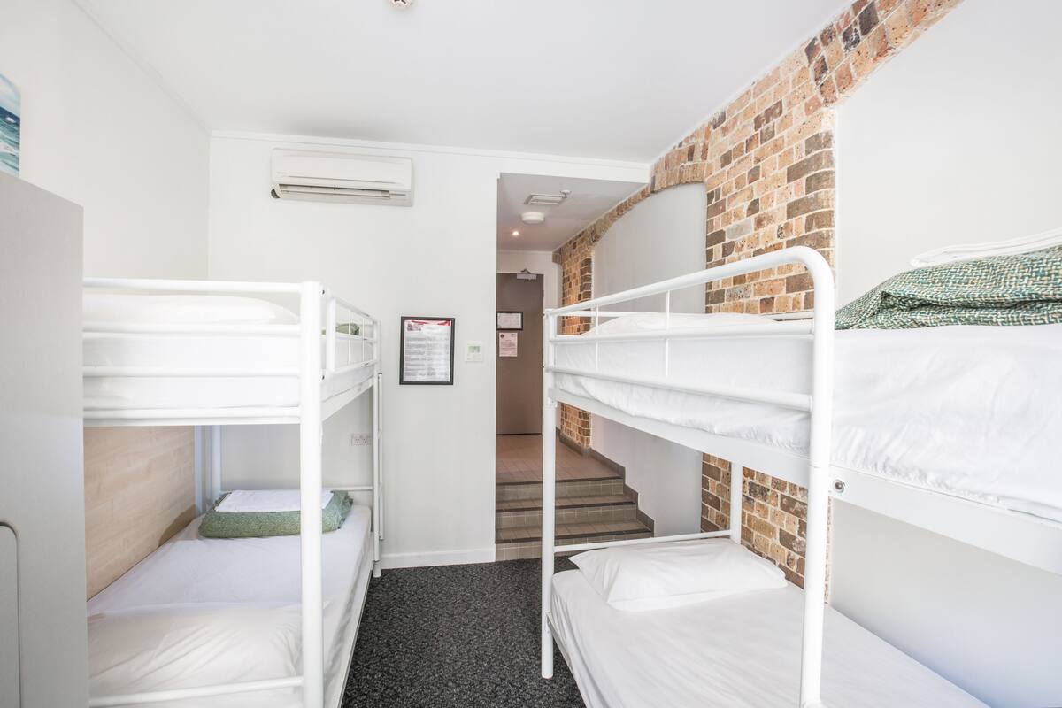 A dormitory room with bunk beds, a brick wall, and a staircase leading to another room.