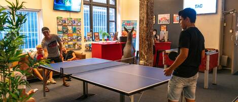 Un tavolo da ping pong in una stanza con una grande finestra, uno schermo televisivo e un poster appeso al muro.