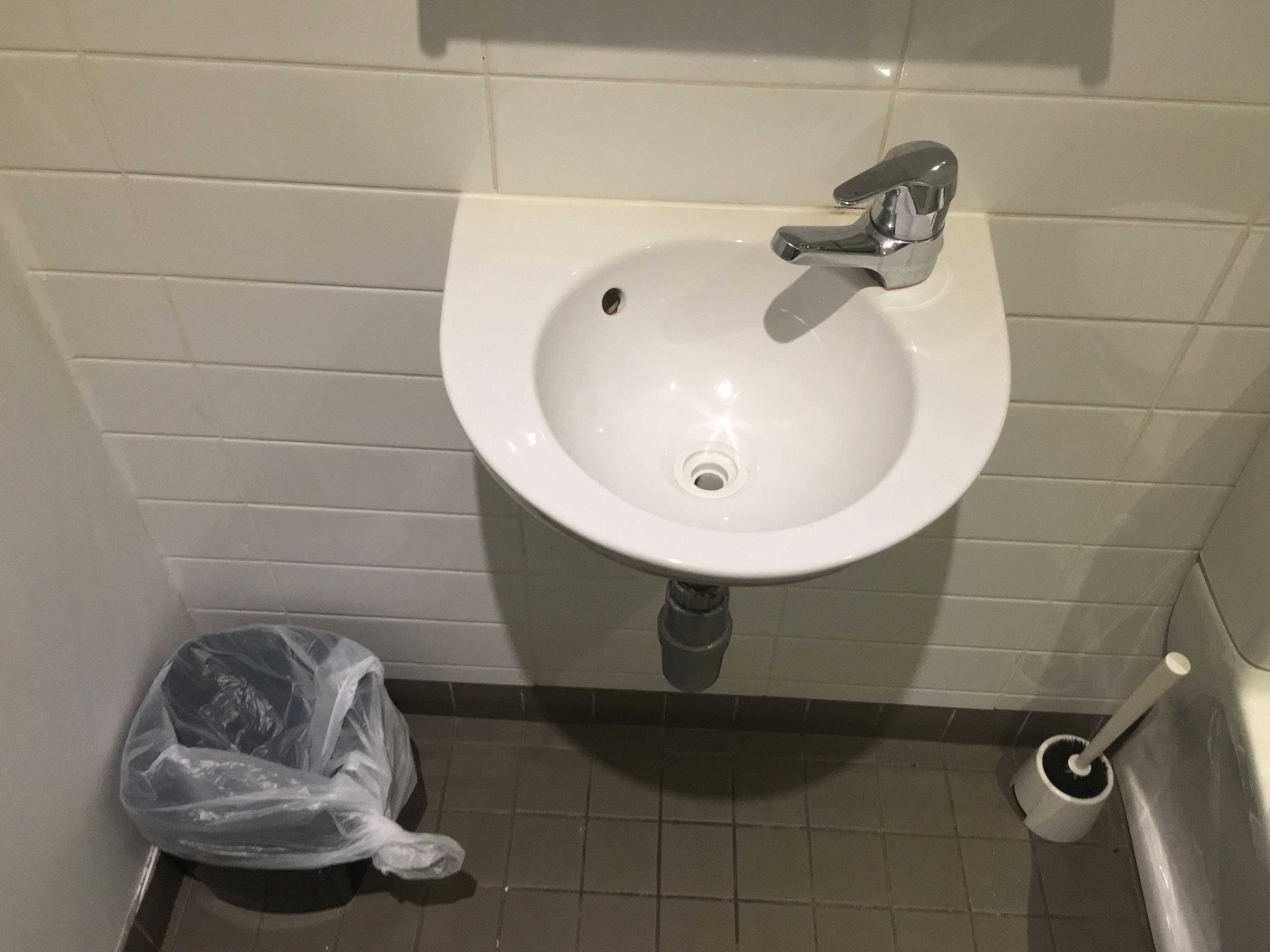 A bathroom sink with a chrome faucet, a plastic trash bag on the floor, and a toilet brush.