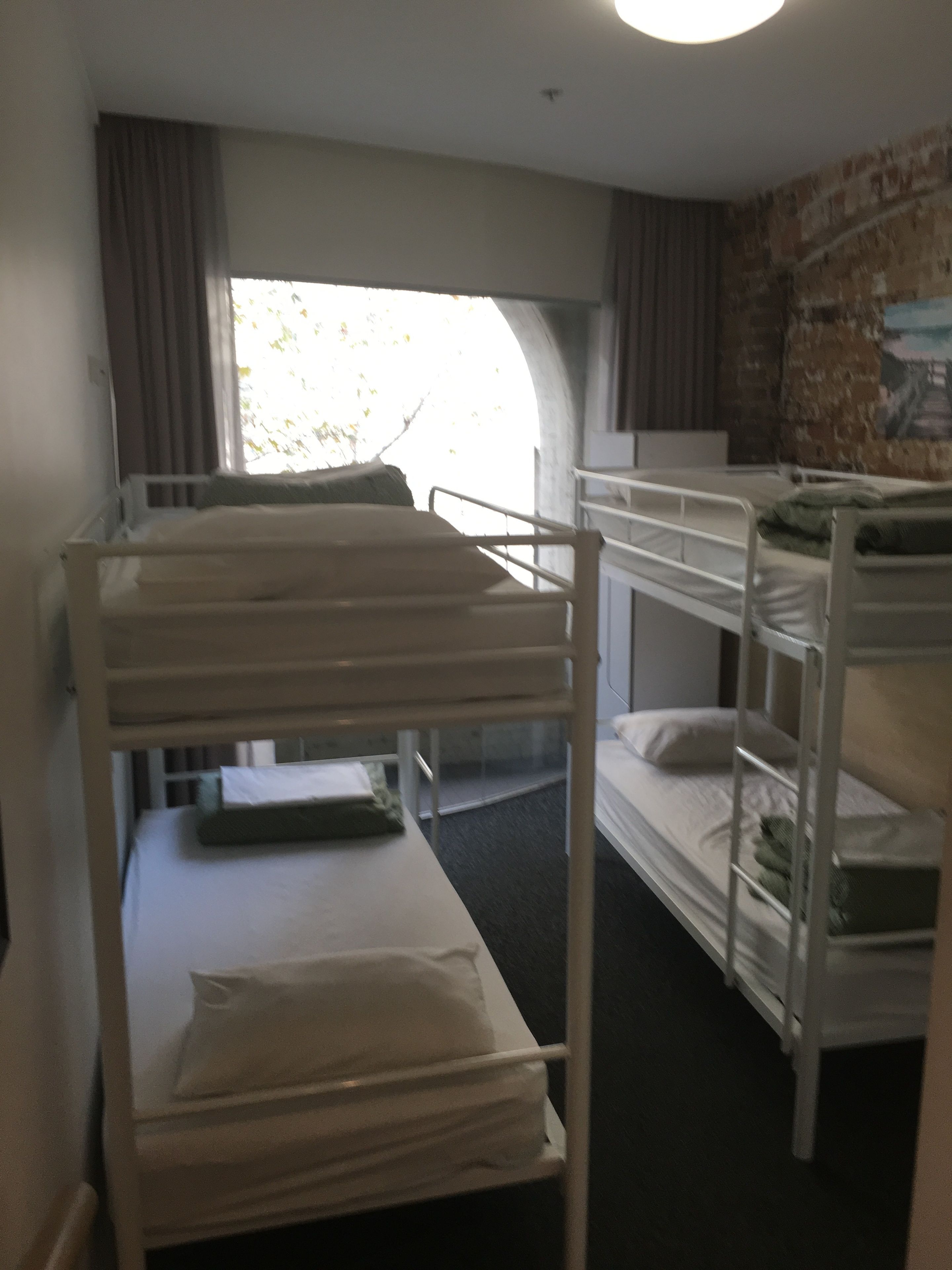 A dormitory room with bunk beds, a window with curtains, and a brick wall with a mural.