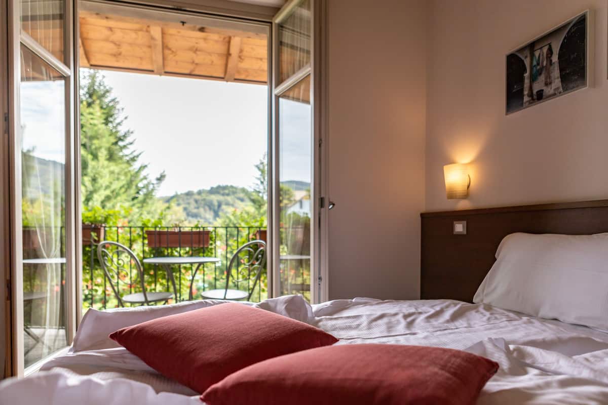 Habitación de hotel con una cama, almohadas rojas, una mesita de noche, una lámpara de pared y un balcón con mesa y sillas.