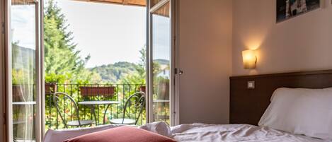A hotel room with a bed, red pillows, a nightstand, a wall-mounted lamp, and a balcony with a table and chairs.
