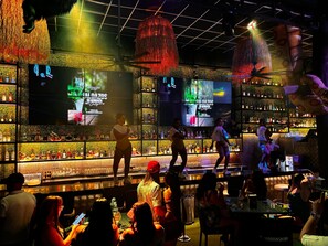 Un bar con techo a rejilla, luces colgantes y una pantalla grande mostrando un anuncio promocional.