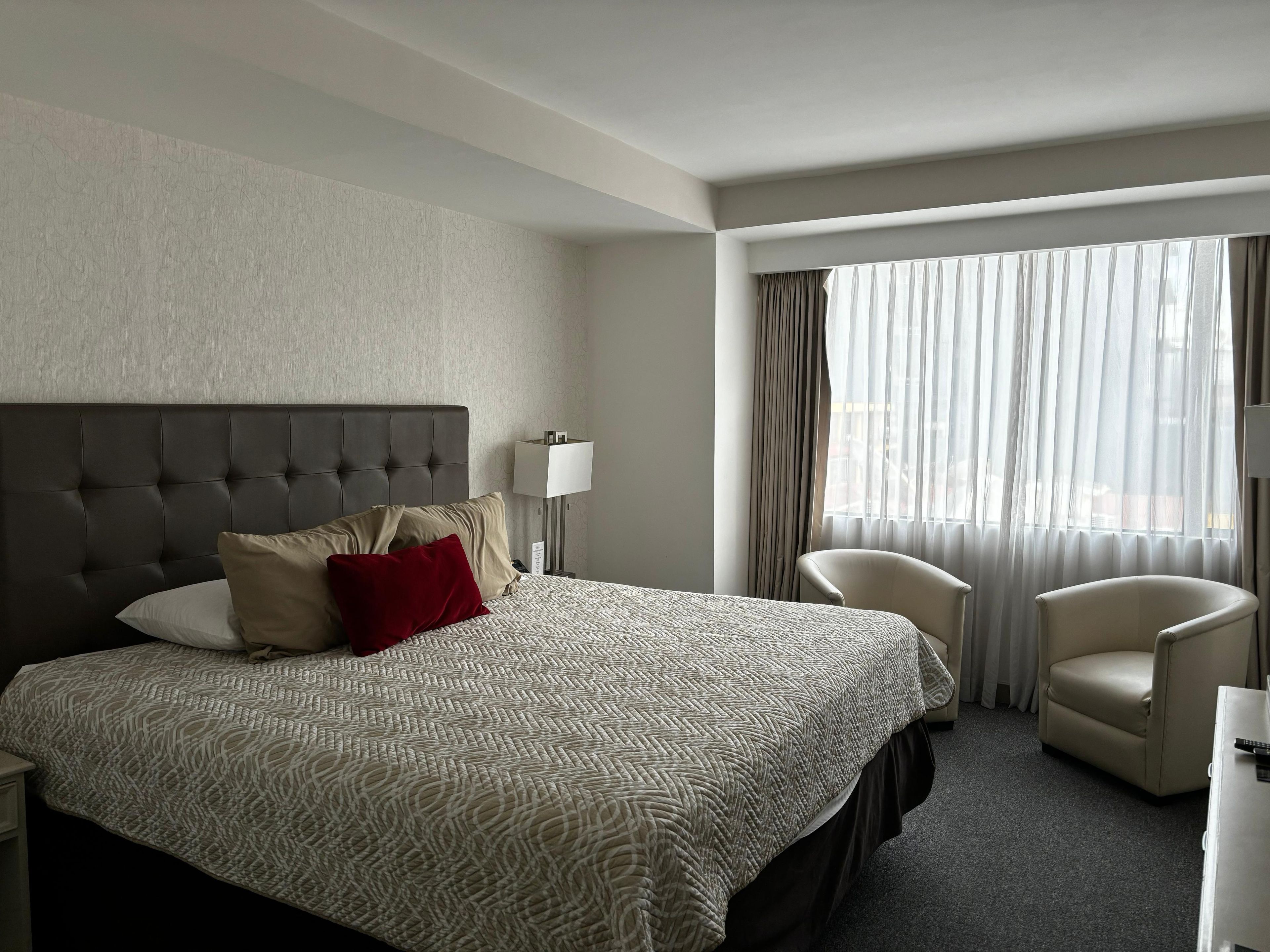 A hotel room with a large bed, a headboard, a bedside table, a lamp, a chair, and a window with curtains.