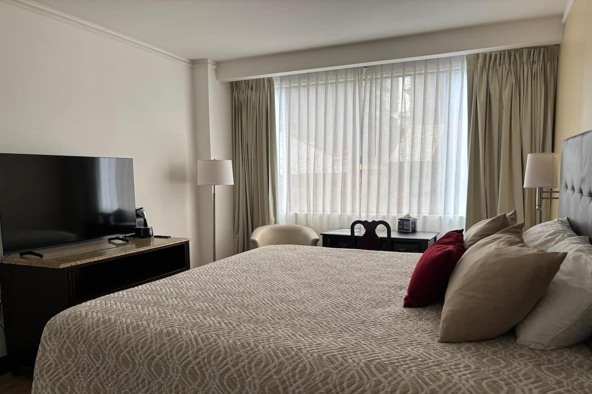 A hotel room with a large bed, a flat-screen TV, a desk with a lamp, a chair, and a view of the city through sheer curtains.