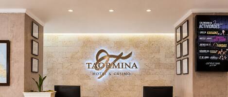 A reception desk with a logo for Taormina Hotel & Casino, framed certificates, and a wall-mounted calendar listing events.