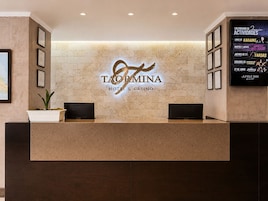 Un mostrador de recepción con el logotipo del Hotel & Casino Taormina, certificados enmarcados y un calendario mural que anuncia eventos.