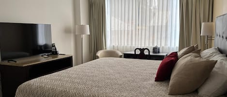 A hotel room with a large bed, a flat-screen TV, a desk with a lamp, a chair, and a view of the city through sheer curtains.
