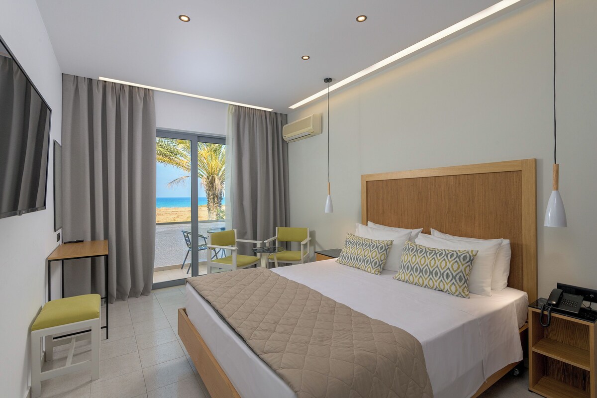 A modern hotel room with a large bed, a desk, a chair, and a view of the beach.