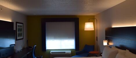 A hotel room with a bed, desk, chair, lamp, and air conditioning unit.