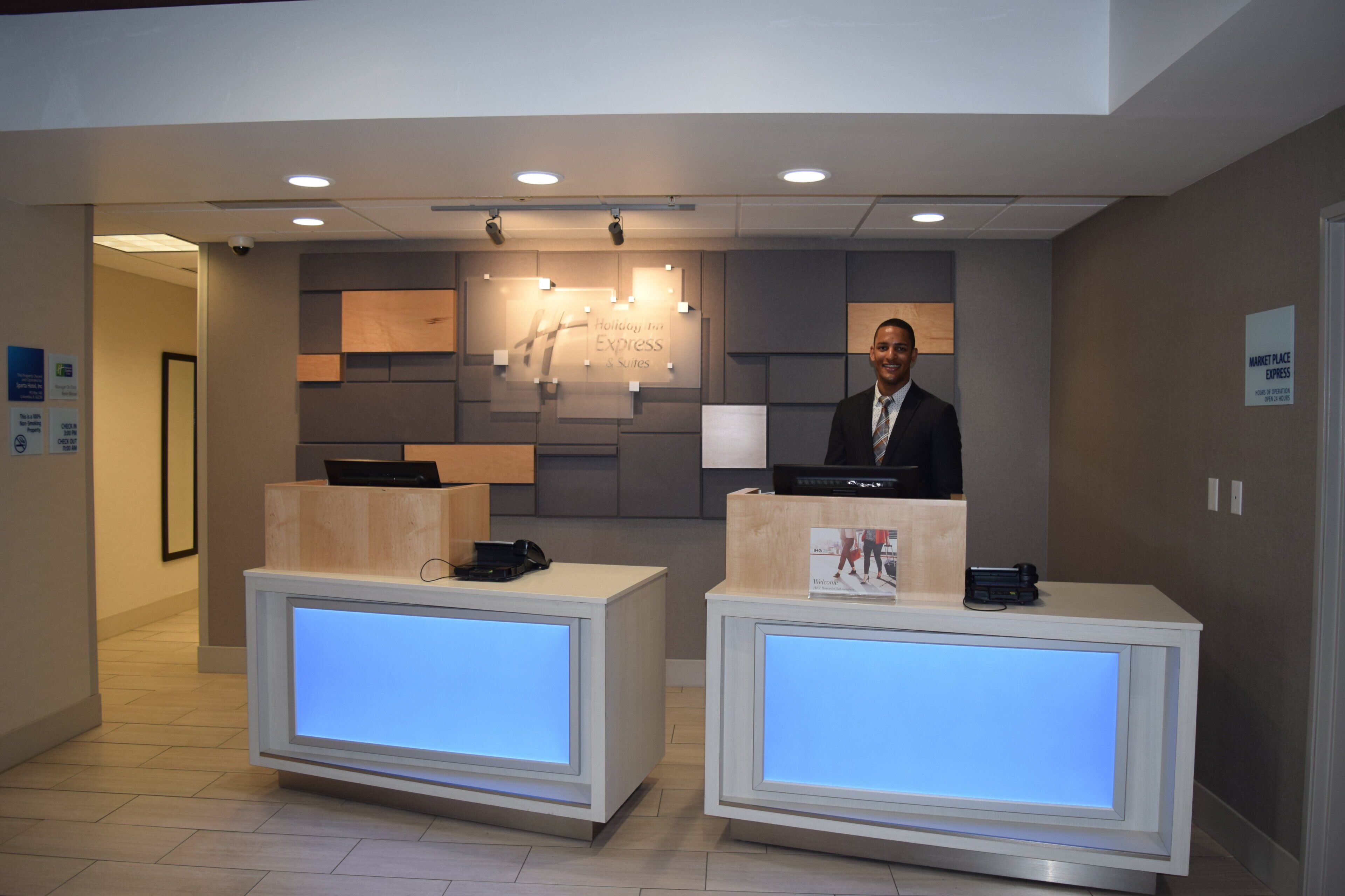 A hotel reception desk with a digital display, a desk phone, and a computer. There is a sign indicating the hotel's name and services.