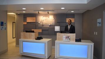 A hotel reception desk with a digital display, a desk phone, and a computer. There is a sign indicating the hotel's name and services.