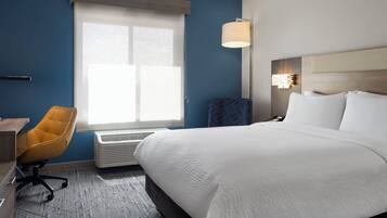 A hotel room with a large bed, a desk with a chair, a window with blinds, and a wall-mounted lamp.