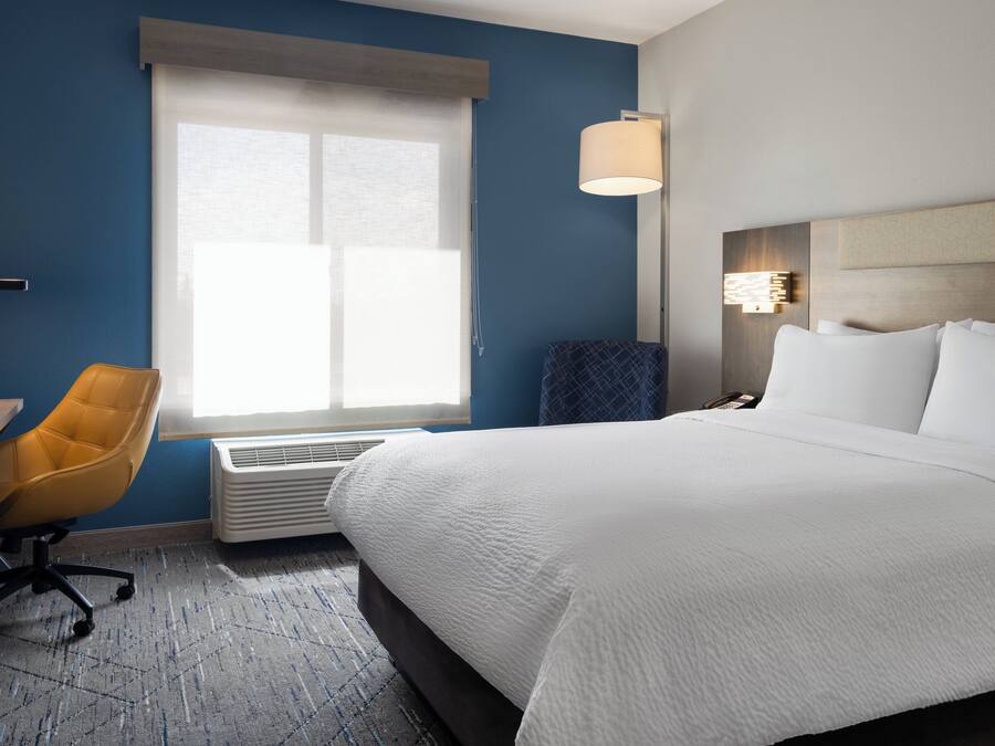 A hotel room with a large bed, a desk with a chair, a window with blinds, and a wall-mounted lamp.