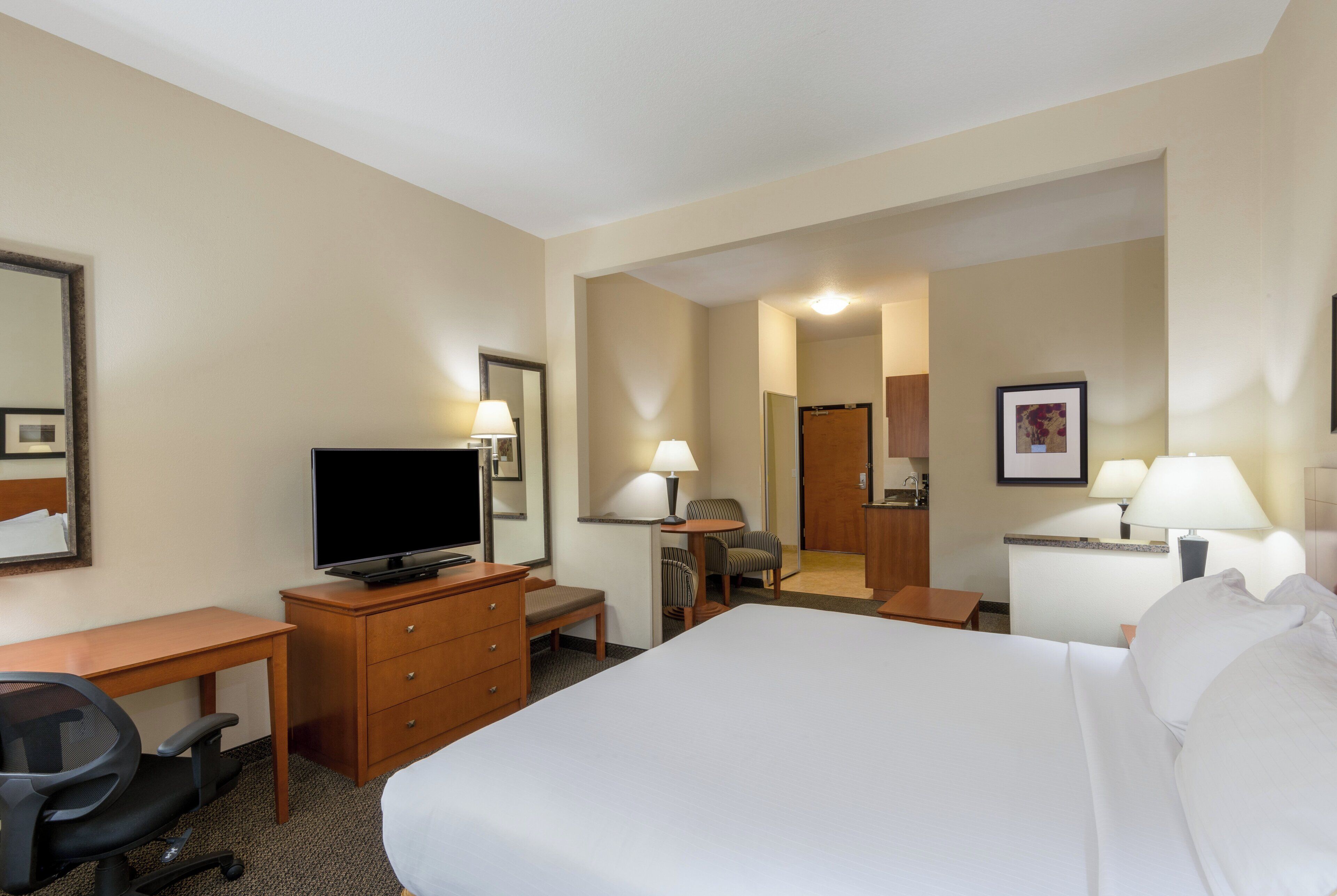 Une chambre d’hôtel avec un lit, un bureau avec une télévision, une chaise, une commode et une salle de bain visible à travers une porte ouverte.