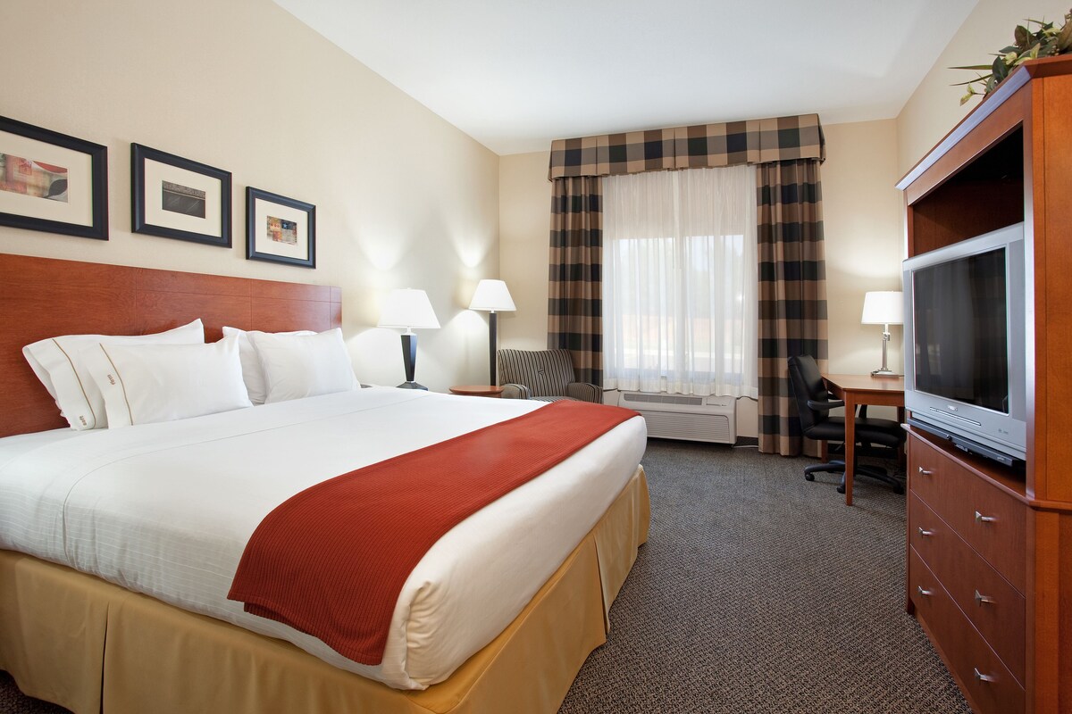 A hotel room with a large bed, two bedside tables with lamps, a television on a wooden cabinet, a desk with a chair, and a window with curtains.