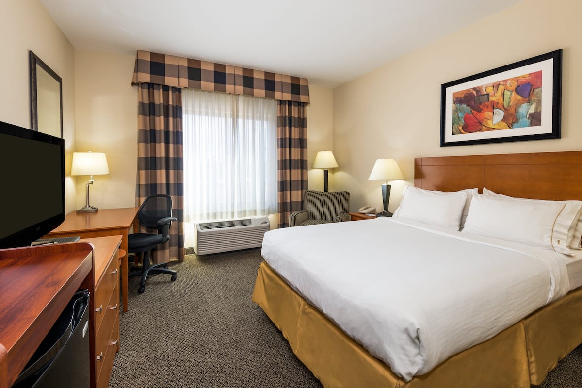 A hotel room with a bed, desk, chair, TV, lamps, and a painting on the wall.