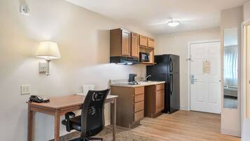 A compact kitchen with wooden cabinets, a microwave, a refrigerator, a small countertop, a chair, a desk, and a lamp.