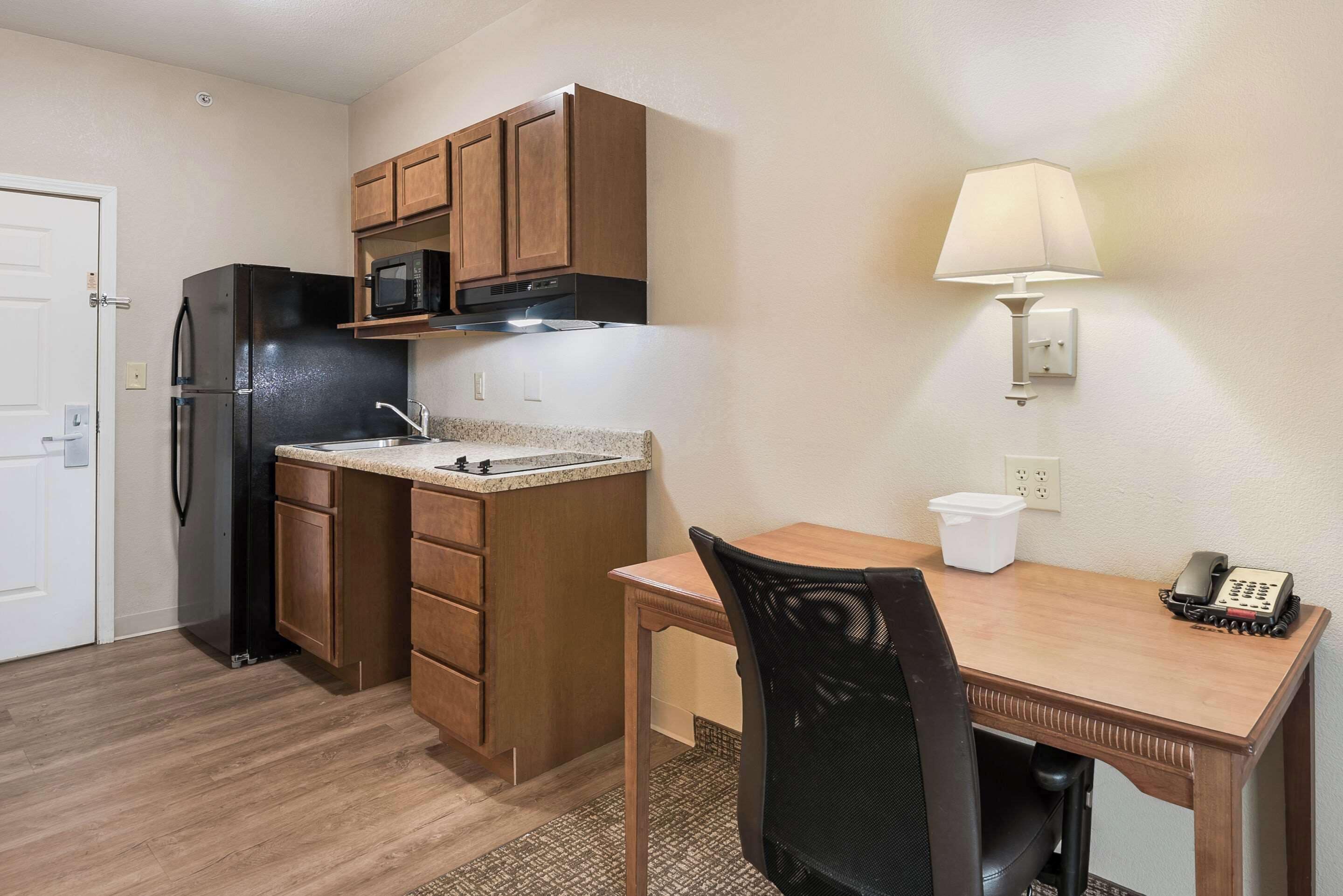 A compact kitchen with wooden cabinets, a black refrigerator, a microwave, a sink, and a small dining table with a chair. There is a wall-mounted lamp and a telephone on the table.