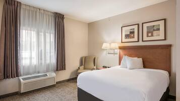 A hotel room with a large bed, a chair, a desk with a lamp, and an air conditioning unit.