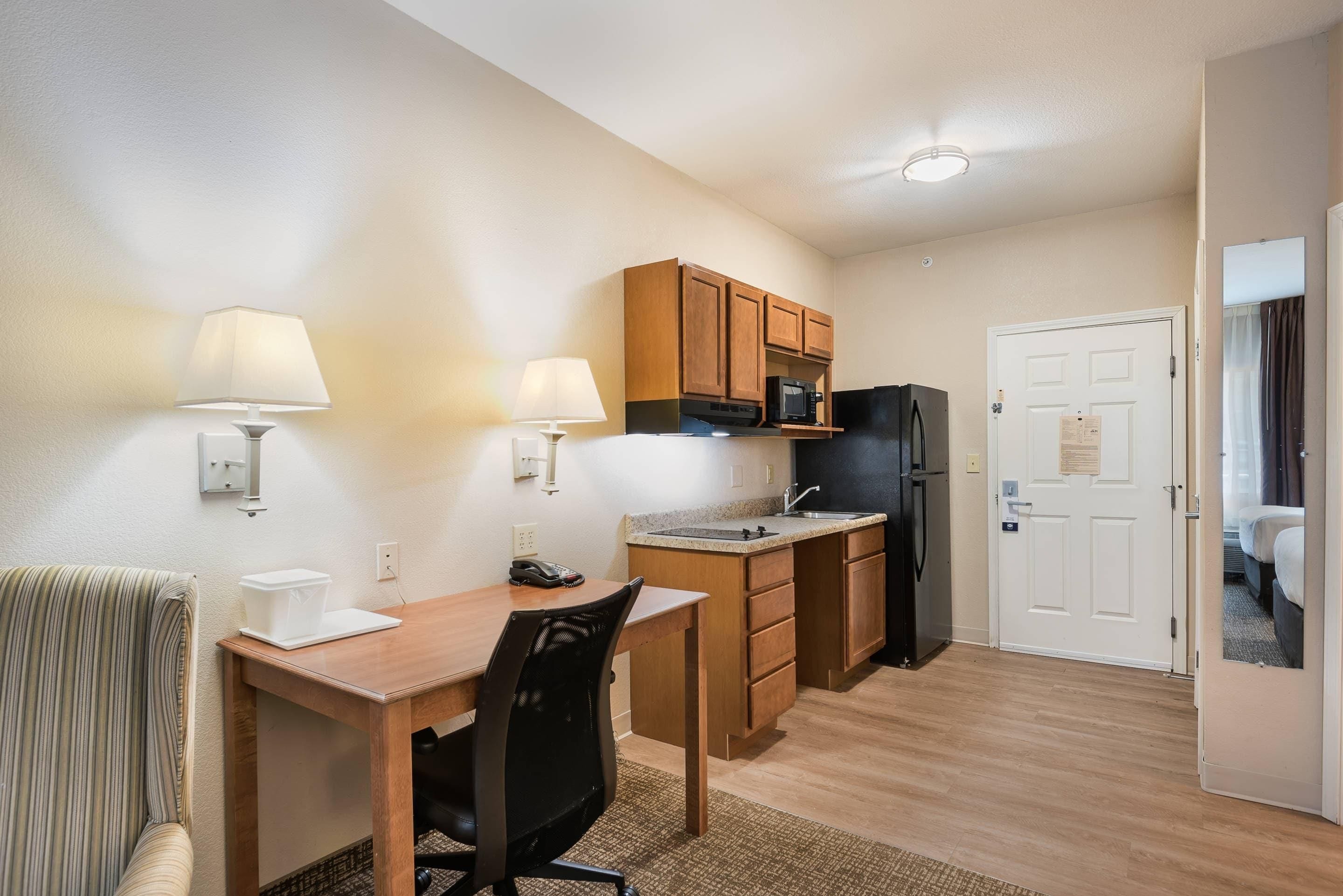 A compact kitchen with wooden cabinets, a refrigerator, a microwave, and a sink. There is a dining table with chairs and two wall-mounted lamps.