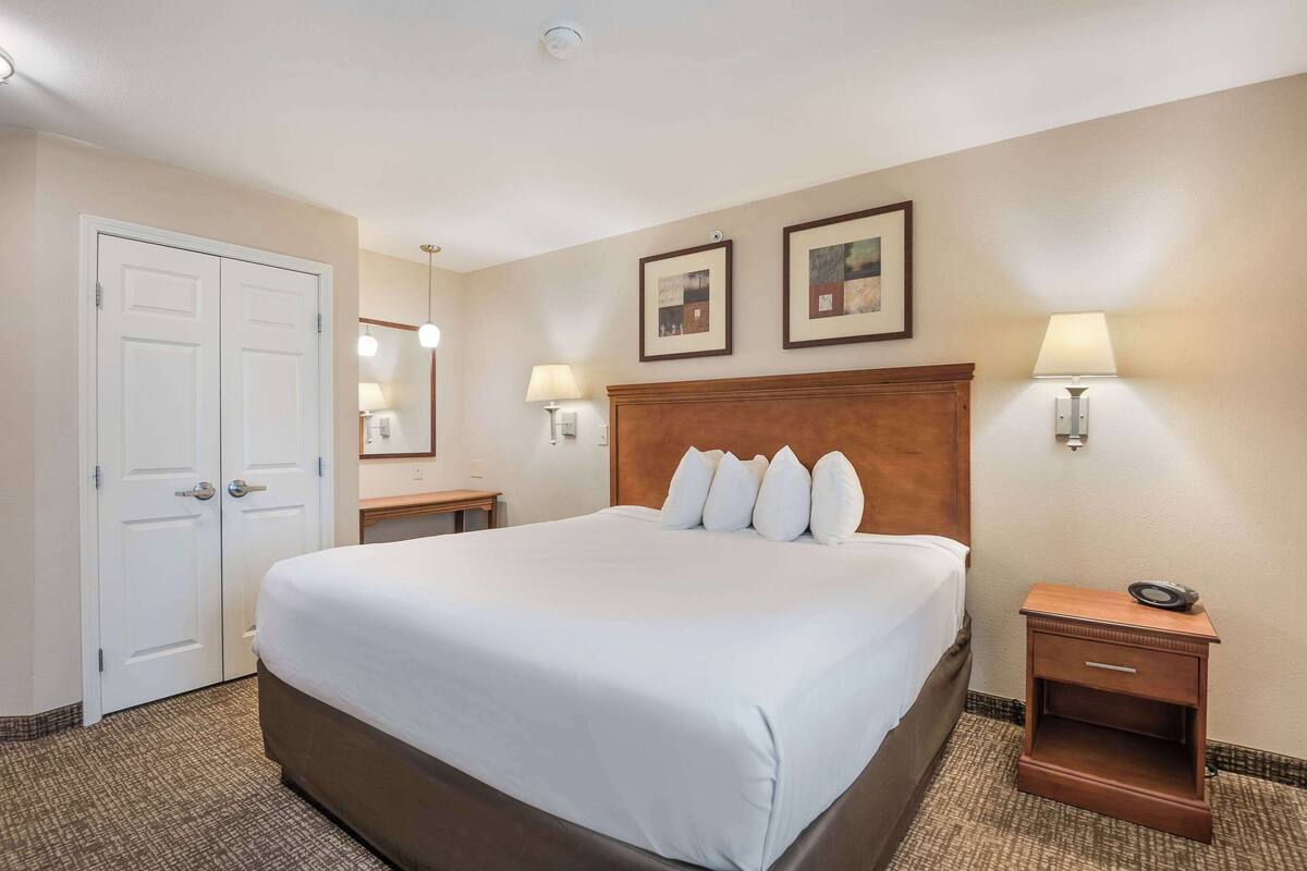 A hotel room with a large bed, a wooden headboard, two bedside lamps, a nightstand, a mirror, and two framed pictures on the wall.