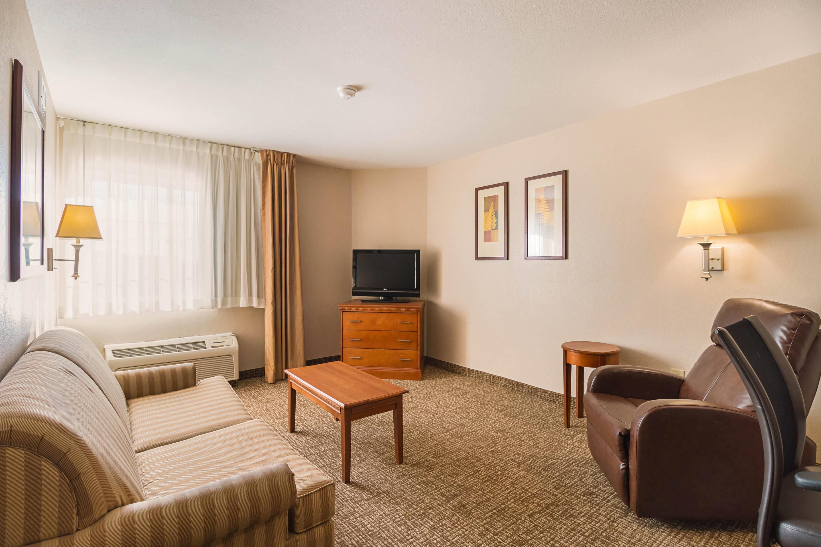 A hotel room with a sofa, a chair, a television on a wooden cabinet, a small table, and two wall-mounted lamps.