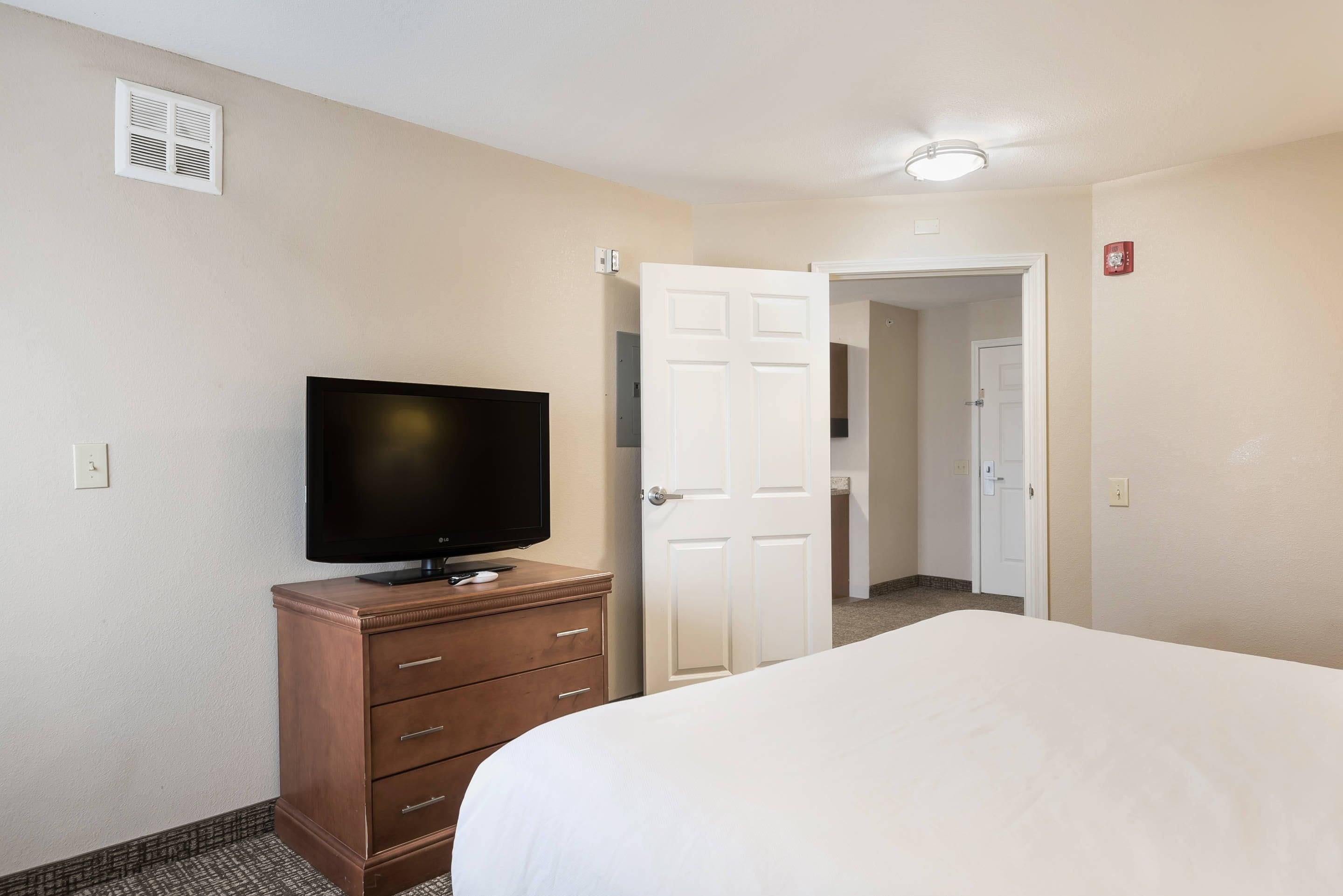 Une chambre d’hôtel avec un lit, un téléviseur posé sur une commode en bois, une porte blanche et un luminaire au plafond.