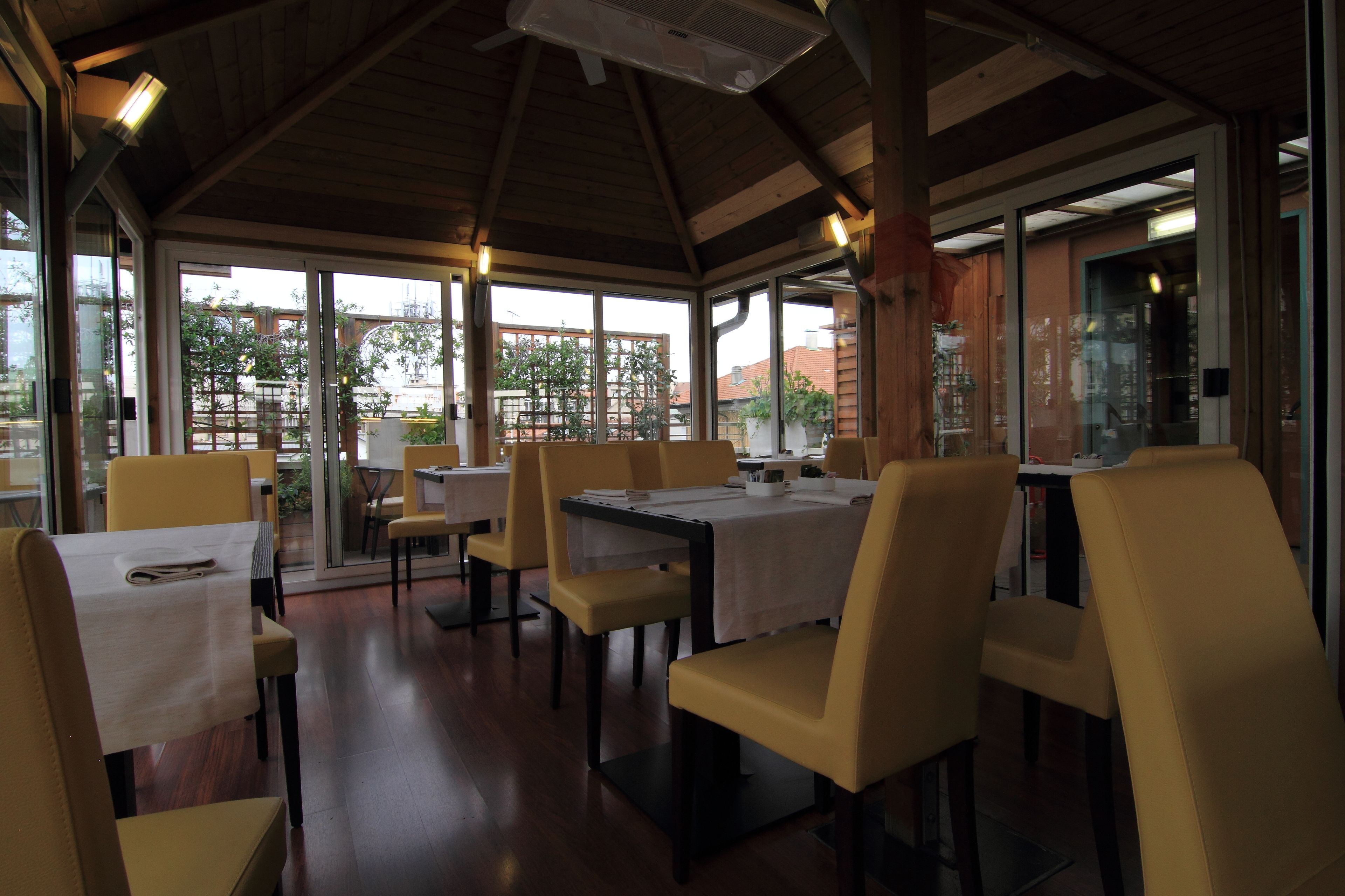 A restaurant with wooden beams, yellow chairs, and tables. Large windows offer views of greenery and buildings.