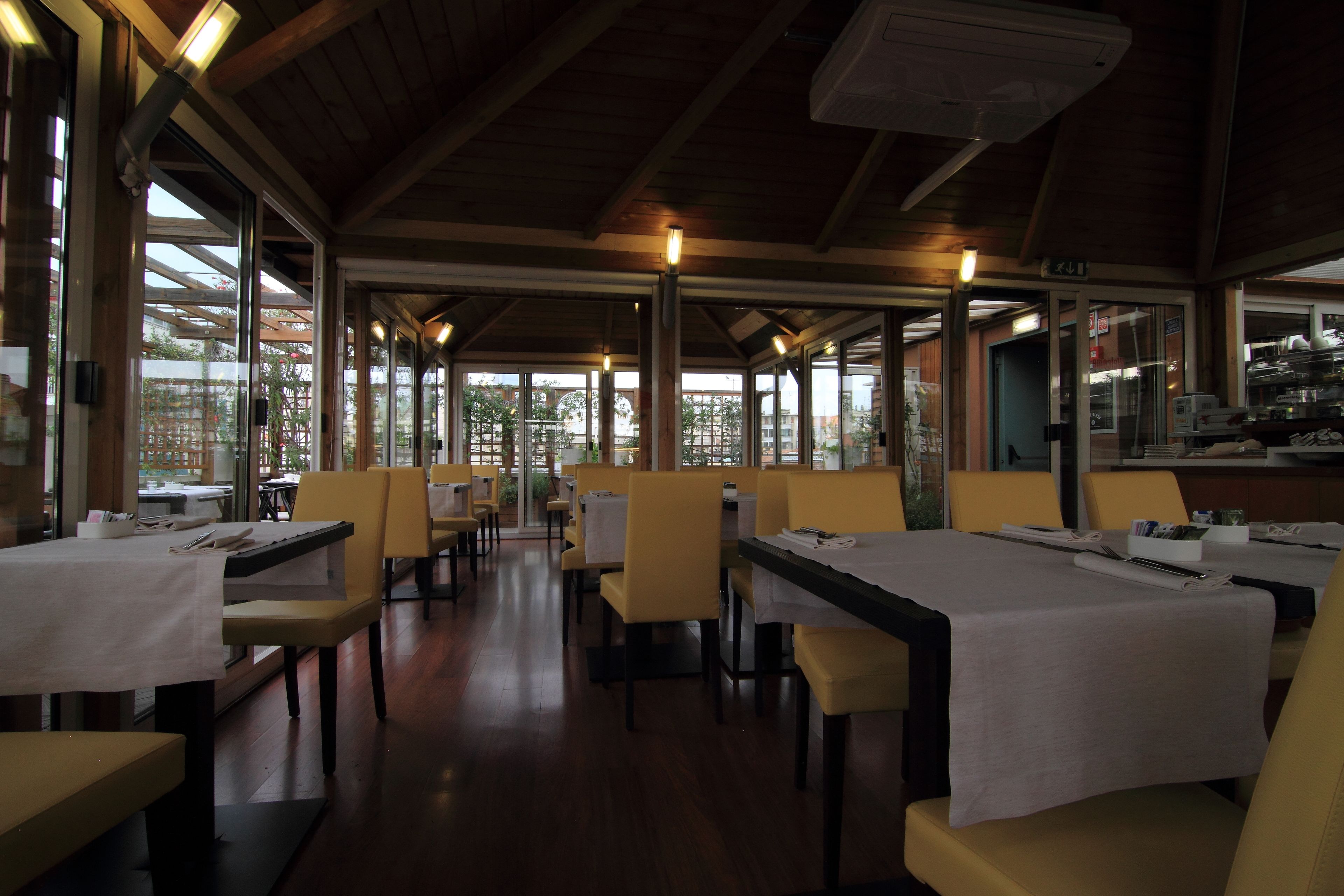 A restaurant with wooden beams, large windows, and yellow chairs. Tables are set with white tablecloths and napkins.