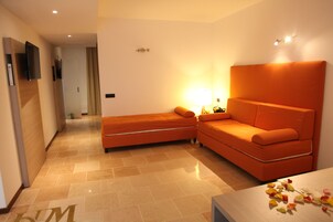 Una camera d'albergo moderna con un ampio divano arancione, un divano più piccolo, una TV a schermo piatto montata a parete e un comodino con una lampada.