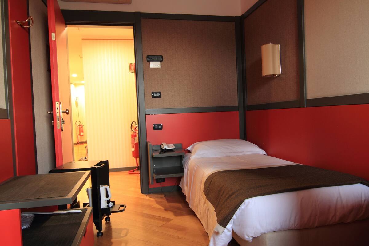 A hotel room with a bed, a desk, and a chair. There is a small table with a chair, a shelf with a phone, and a wall-mounted light.
