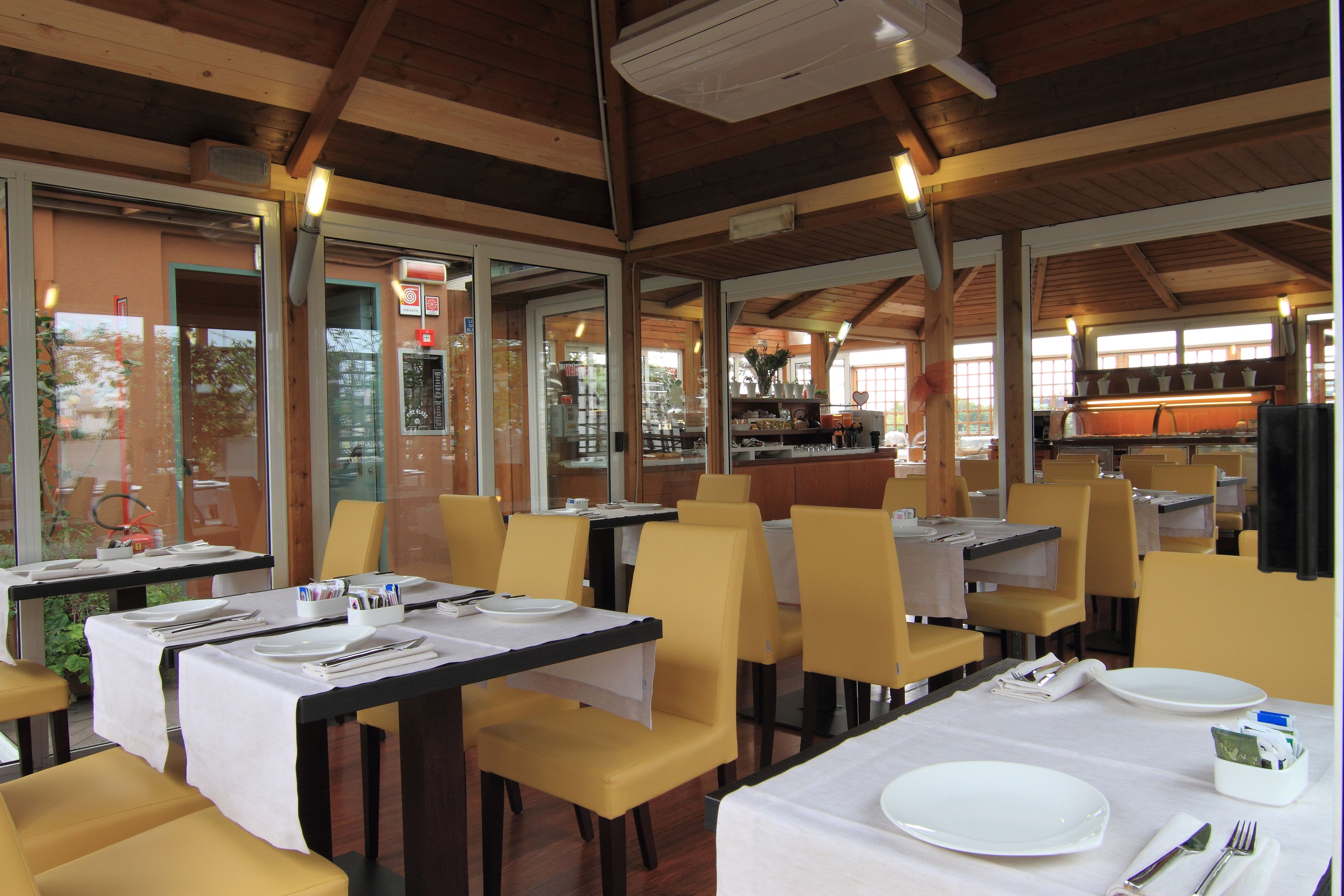 A restaurant with tables set for dining, yellow chairs, and a kitchen area visible through the glass doors.