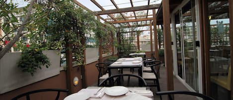 An outdoor dining area with tables and chairs, covered by a wooden pergola with glass panels.